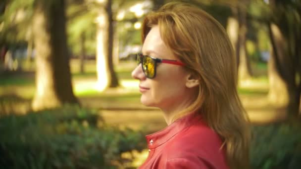 Una mujer con gafas de sol y una chaqueta roja pasea por el parque de la ciudad en un día claro y soleado. 4k, cámara lenta — Vídeos de Stock