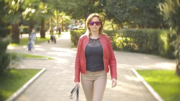 Uma mulher de óculos de sol e um casaco vermelho passeia pelo parque da cidade em um dia ensolarado claro. 4k, câmera lenta — Vídeo de Stock