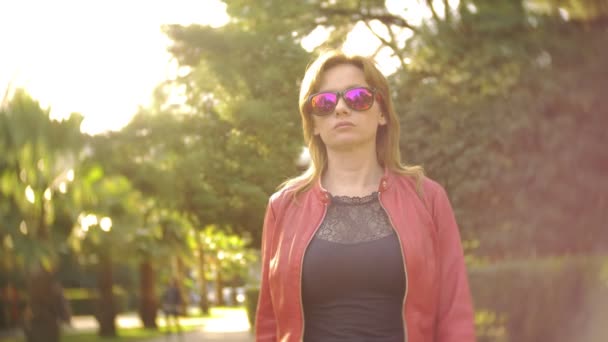 Una mujer con gafas de sol y una chaqueta roja pasea por el parque de la ciudad en un día claro y soleado. 4k, cámara lenta — Vídeos de Stock