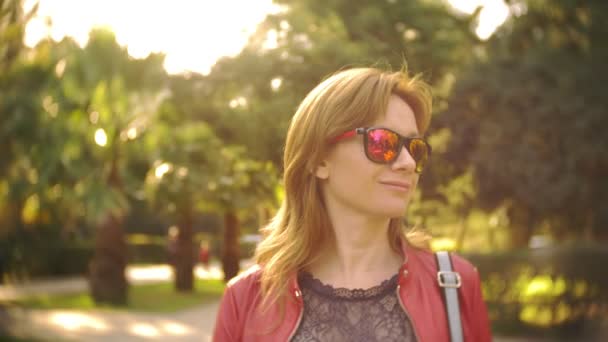 Una mujer con gafas de sol y una chaqueta roja pasea por el parque de la ciudad en un día claro y soleado. 4k, cámara lenta — Vídeos de Stock