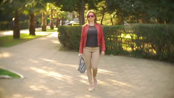 Egy nő a napszemüveg és a piros kabát sétál átmenő a városi park, egy világos napsütéses napon. 4k, lassú mozgás — Stock videók