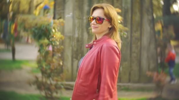 Une femme portant des lunettes de soleil et une veste rouge se promène dans le parc de la ville par une journée ensoleillée. 4k, au ralenti — Video