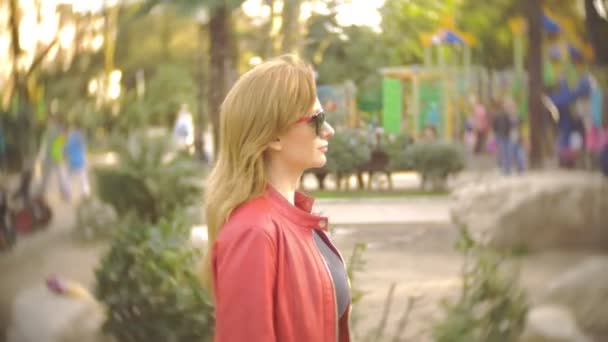 Uma mulher de óculos de sol e um casaco vermelho passeia pelo parque da cidade em um dia ensolarado claro. 4k, câmera lenta — Vídeo de Stock