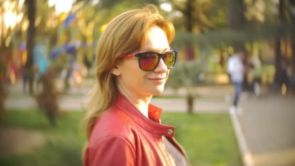 Žena v sluneční brýle a červenou bundu procházek přes městský park na jasný slunečný den. 4k, pomalý pohyb — Stock video