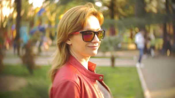 Une femme portant des lunettes de soleil et une veste rouge se promène dans le parc de la ville par une journée ensoleillée. 4k, au ralenti — Video