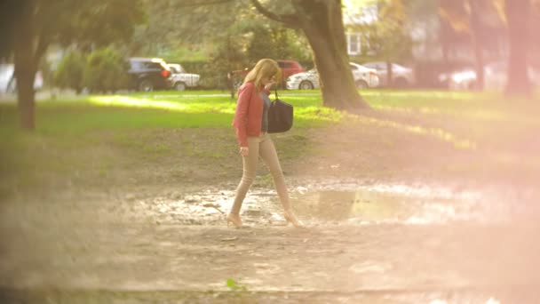 Тонкая женщина в бежевых брюках и туфлях на высоких каблуках, прогуливается по парку мимо луж на лужайке. 4k, slow motion — стоковое видео