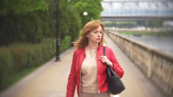 A woman with long hair in a red leather jacket walks along the city embankment. 4k, background blur — Stock Video