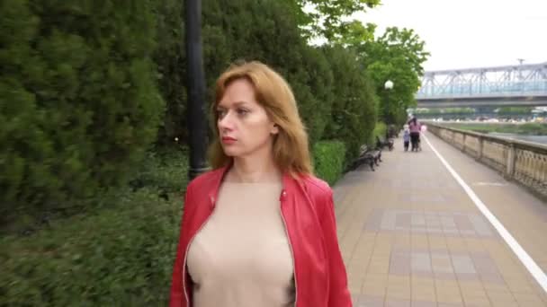 Une femme aux cheveux longs dans une veste en cuir rouge se promène le long du remblai de la ville. 4k, flou de fond — Video