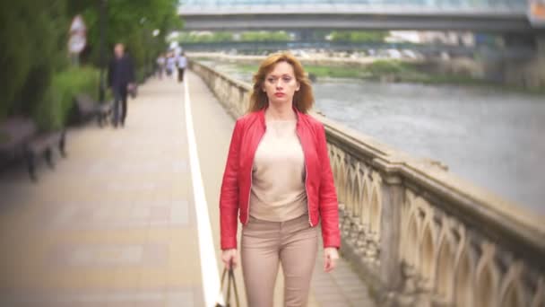 По городской набережной ходит женщина с длинными волосами в красной кожаной куртке. 4K, размытие фона — стоковое видео