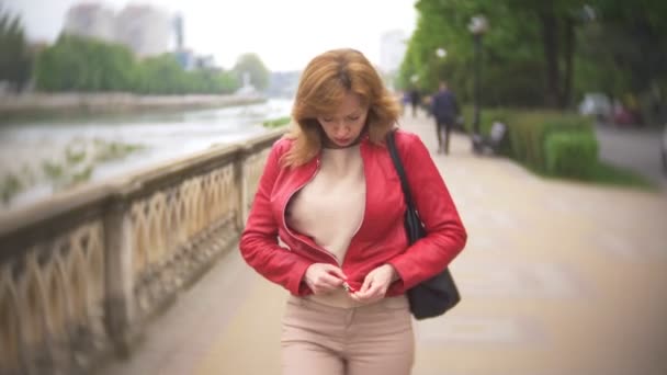 Una mujer con el pelo largo en una chaqueta de cuero rojo camina a lo largo del terraplén de la ciudad. 4k, desenfoque de fondo — Vídeo de stock
