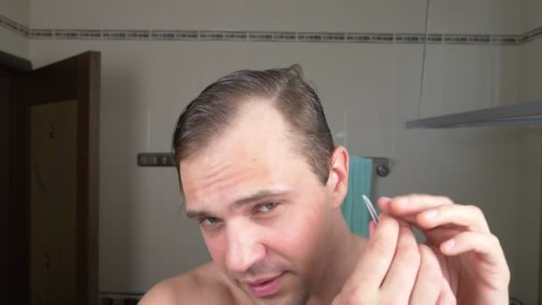 Un bell'uomo tira fuori i capelli dalle sopracciglia con delle pinzette davanti a uno specchio in bagno. Epilazione delle sopracciglia. 4k — Video Stock