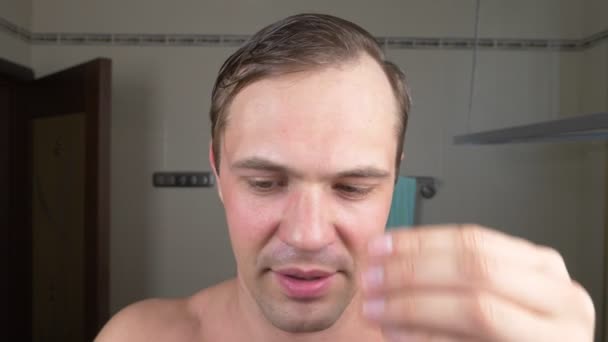 Een knappe man trekt zijn haar uit zijn wenkbrauwen met pincet voor een spiegel in de badkamer. Epileren van de wenkbrauw. 4k — Stockvideo