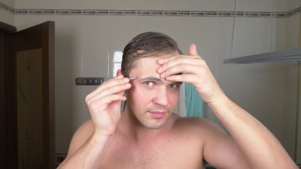 Un bel homme sort ses cheveux de ses sourcils avec une pince à épiler devant un miroir dans la salle de bain. Épilation des sourcils. 4k — Video