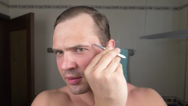En stilig man drar sitt hår ur ögonbrynen med en pincett framför en spegel i badrummet. Ögonbryn epilering. 4k — Stockvideo