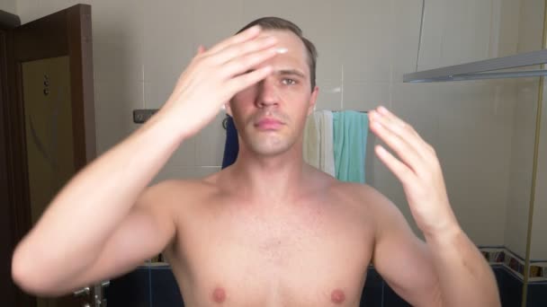 Um homem bonito mancha um rosto com um creme na frente de um espelho na casa de banho. 4k — Vídeo de Stock