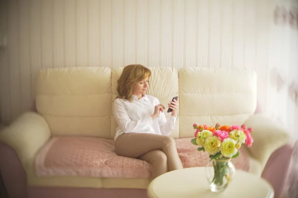 Een trendy dame gekleed in een witte blouse en een beige broek. Een jonge vrouw maakt gebruik van een mobiele telefoon binnenshuis met een prachtige interieur. — Stockfoto