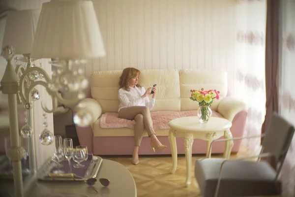 Een trendy dame gekleed in een witte blouse en een beige broek. Een jonge vrouw maakt gebruik van een mobiele telefoon binnenshuis met een prachtige interieur. — Stockfoto