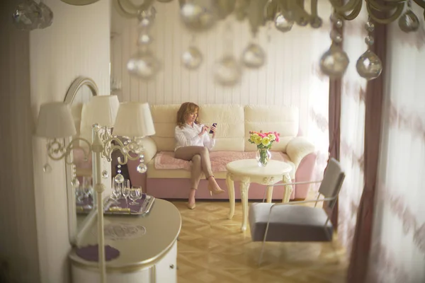 A fashionable lady dressed in a white blouse and beige trousers. A young woman uses a mobile phone indoors with an exquisite interior. — Stock Photo, Image