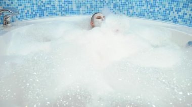 Yüzünde maske olan kel genç bir kadın köpükle banyoda yatıyor, gülümsüyor ve köpüğe üflüyor.