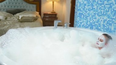 Yüzünde maske olan kel genç bir kadın köpükle banyoda yatıyor, gülümsüyor ve köpüğe üflüyor.