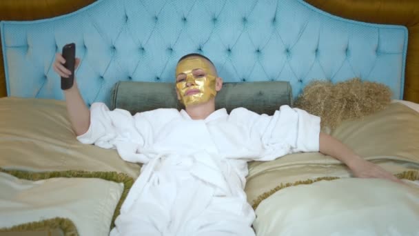 Yüzünde altın bir maske olan genç sarışın kadın, televizyon seyrediyor, altın bir yatağın üzerinde yatıyor, peruğunu çıkarıyor ve kel olduğu ortaya çıkıyor. — Stok video