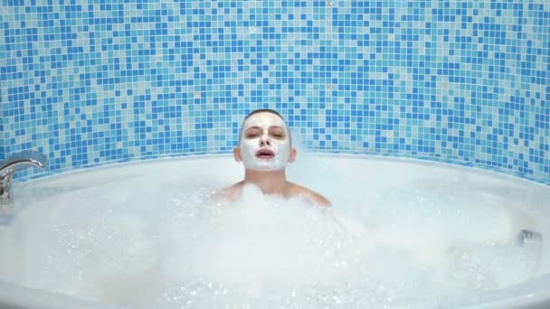 A bald young woman with a mask on her face lies in a bath with foam, she smiles and blows on the foam — Stock Video