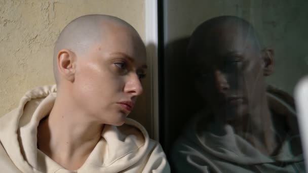 A sad bald woman sits on a windowsill by the window at night and looks out the window. reflection in glass — Stock Video