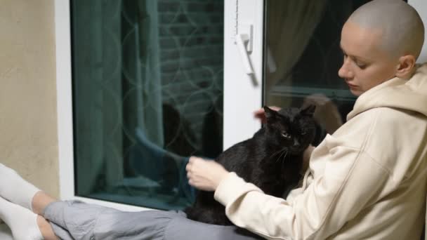 Triste mujer calva se sienta en la noche en un alféizar de la ventana y acaricia a un gato negro — Vídeo de stock