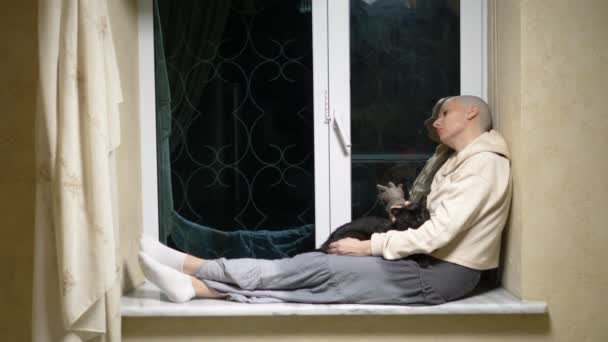 Triste mujer calva se sienta en la noche en un alféizar de la ventana y acaricia a un gato negro — Vídeo de stock