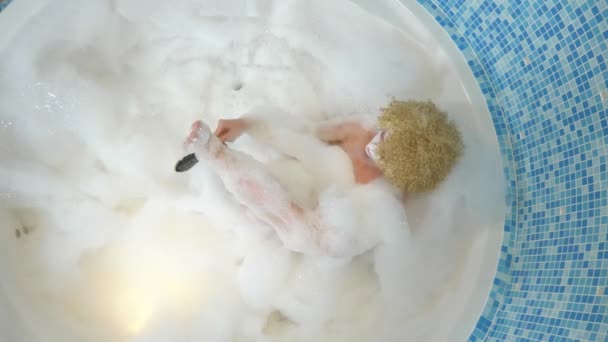 Joven mujer hermosa en un baño de lujo con espuma gruesa pule la piel del pie con piedra pómez. Concepto de cuidado corporal. vista desde arriba — Vídeo de stock