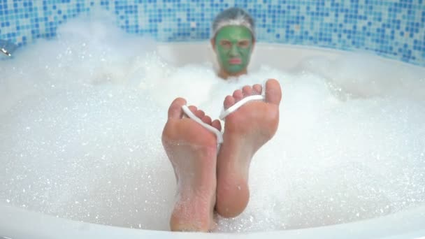 A young man in a shower cap and with a green cosmetic mask on his face lies in the bathroom with copious foam. his feet with finger dividers are visible from the water, in focus — 비디오