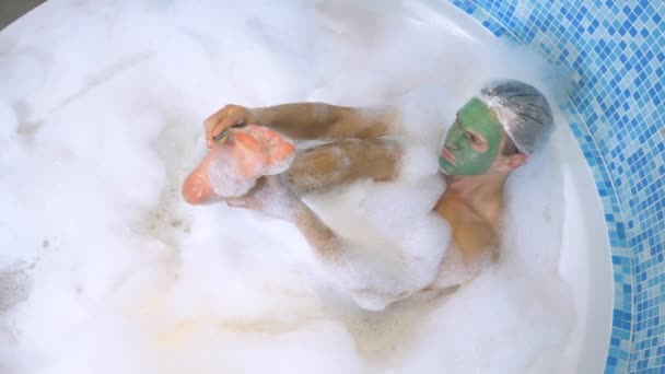 Un uomo con una cuffia da doccia e una maschera verde si trova in un bagno con abbondante schiuma e si lava il piede con la pomice. igiene con scrub nel bagno. vista dall'alto — Video Stock