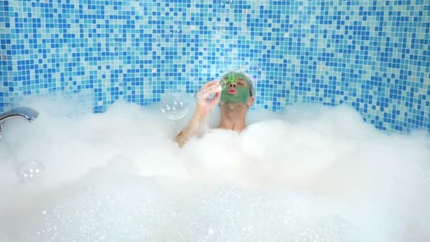 Hombre lindo emocional con máscara de arcilla en una gorra de baño, soplando burbujas de jabón mientras está acostado en una bañera con abundante espuma. concepto humorístico. espacio de copia — Vídeo de stock