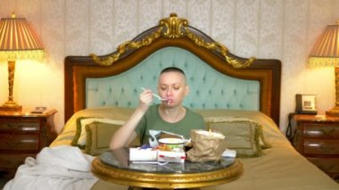 Haki gömlekli, aç ve kel bir kız. Fast food yiyor. Lüks bir yerde, yatağın üzerinde oturuyor.