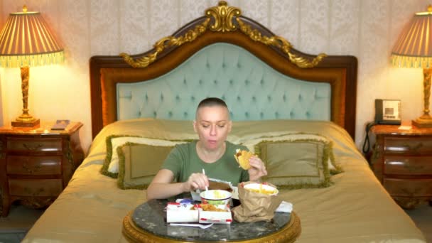 Hungrig skallig flicka i en khaki skjorta äta snabbmat sitter i en lyxig inredning på sängen — Stockvideo