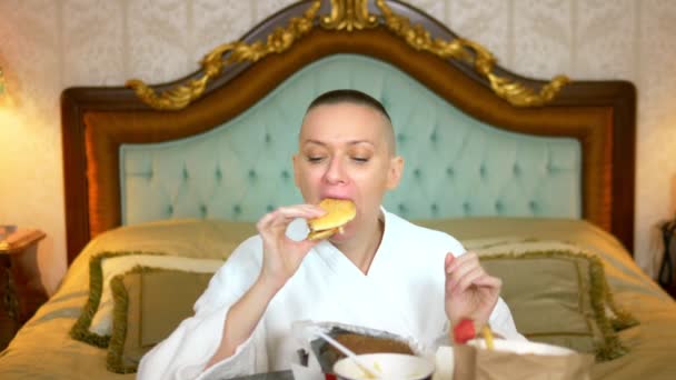 Éhes kopasz lány fürdőköpenyben gyorskaját eszik egy fényűző belső térben az ágyon. — Stock videók