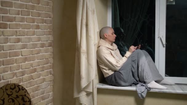 Sad bald woman sits on a windowsill by the window at night and uses the phone — Stock Video