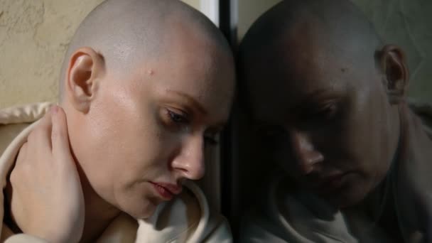 A sad bald woman sits on a windowsill by the window at night and looks out the window. reflection in glass — Stock Video