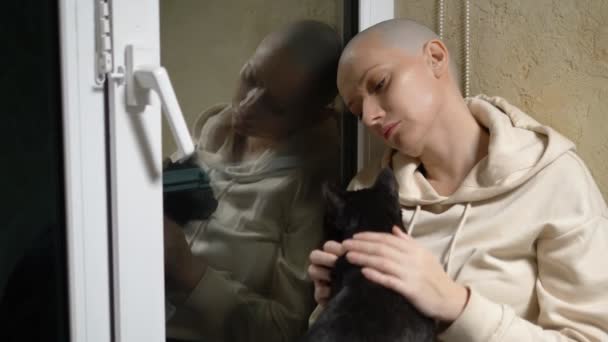 Eine traurige Frau mit Glatze sitzt nachts auf einem Fenstersims am Fenster und streichelt eine schwarze Katze — Stockvideo