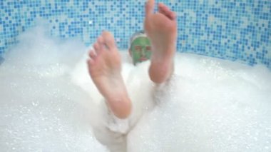 Yüzünde yeşil kozmetik maske olan duş bonesi takmış genç bir adam bol köpükle banyoda yatıyor. Ayakları parmak ayırıcı ile sudan görülebiliyor, odaklanabiliyor.