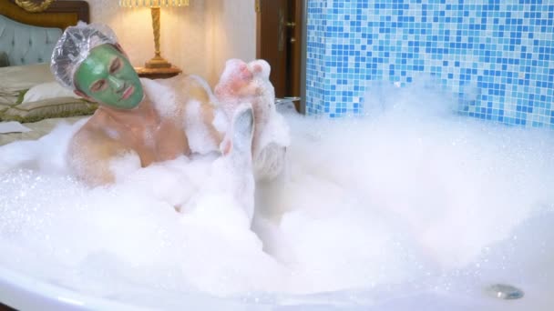 Egy férfi zuhanysapkában és zöld arcmaszkban fekszik a fürdőkádban bőséges habbal, és habszivaccsal mossa a lábát. higiénia mosakodással a kádban. felülnézetből — Stock videók