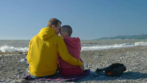 Пара влюбленных мужчина и лысая женщина сидят на берегу моря в прохладную ветреную погоду . — стоковое фото
