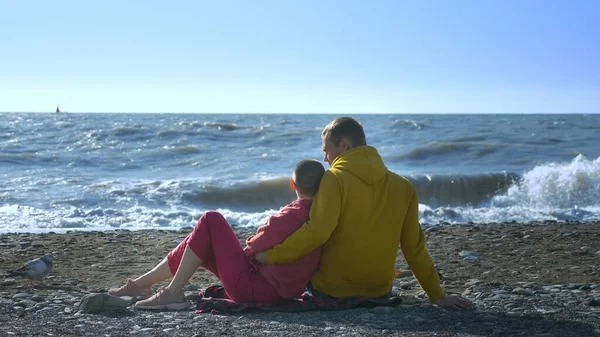 Couple en amour homme et une femme chauve sont assis sur le bord de la mer par temps frais et venteux . — Photo