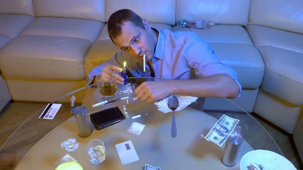 Un hombre en una camisa de oficina drogado en una mesa con botellas de alcohol, cigarrillos, polvo blanco y jeringas . — Foto de Stock