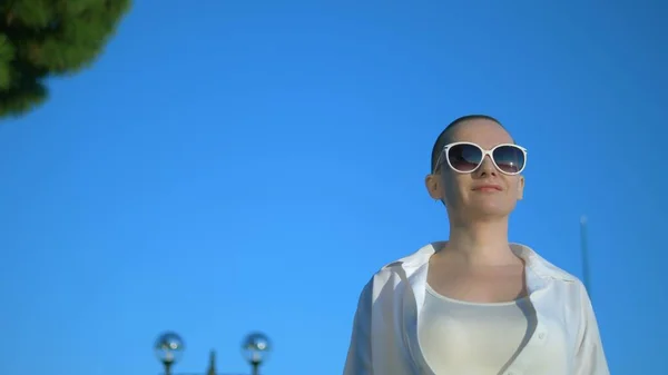 Stilvolles glatzköpfiges Mädchen mit Sonnenbrille und weißem Hemd geht die Straße vor blauem Himmel und grünen Bäumen entlang — Stockfoto