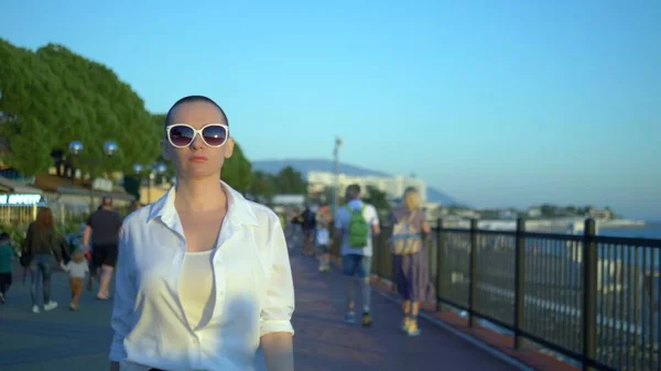 Stilvolles glatzköpfiges Mädchen mit Sonnenbrille und weißem Hemd geht die Straße vor blauem Himmel und grünen Bäumen entlang — Stockfoto