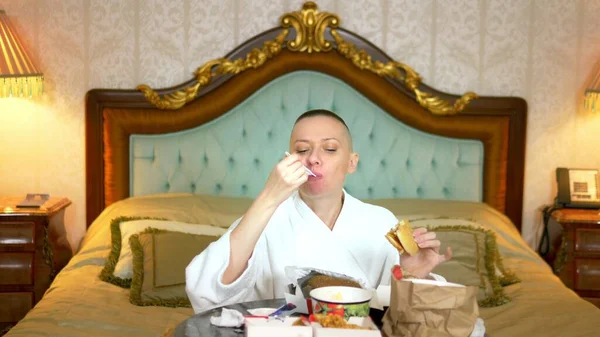 hungry bald girl in a bathrobe eats fast food sitting in a luxurious interior on the bed