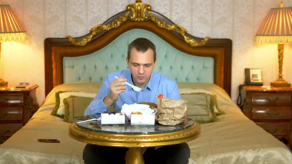 Primer plano. Hombre de negocios guapo está comiendo comida rápida sentado en la cama en un interior de lujo . — Foto de Stock