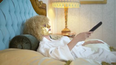 Yüzünde altın bir maske olan genç sarışın kadın altın bir yatakta uzanmış televizyon seyrediyor..