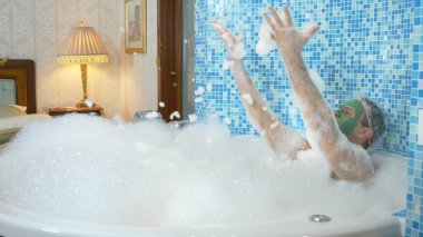 Yüzünde yeşil bir maske ve duş bonesi olan genç bir adam köpükle banyo yapıyor. Adam banyoda dans ediyor, vücudu kalın beyaz köpükle kaplı. Boşluğu kopyala